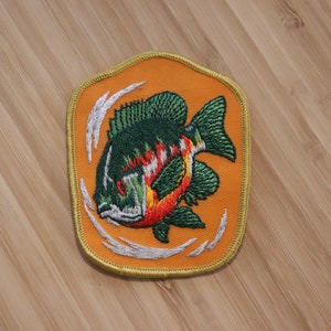 Peut inclure: Un patch brodé orange avec un poisson vert, rouge et jaune nageant dans un cercle de vagues blanches.