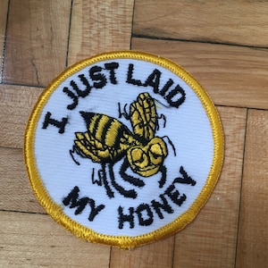 May include: A round, white patch with a yellow border and the text "I JUST LAID MY HONEY". The patch features a cartoon bee with yellow and black stripes and black legs.