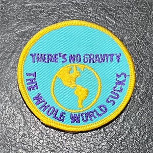 May include: A yellow and blue embroidered patch with the text "There's no gravity the whole world sucks". The patch features a blue globe with a yellow outline.