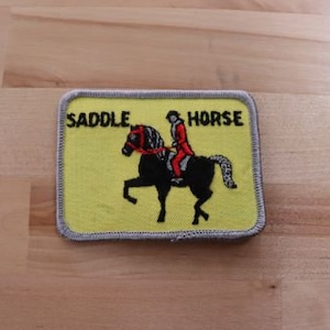 May include: A yellow embroidered patch featuring a black horse and rider. The patch reads "Saddle Horse" in black text.