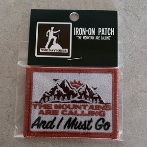 Puede incluir: Un parche termoadhesivo rectangular con borde marrón, con el texto "THE MOUNTAINS ARE CALLING And I Must Go" y un diseño de montaña. El parche está empaquetado con un respaldo negro que dice "IRON-ON PATCH".