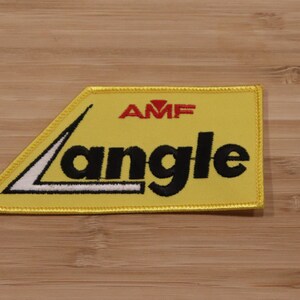 Può includere: Toppa in tessuto giallo con cuciture nere e il testo "AMF" in rosso e "Langle" in nero.