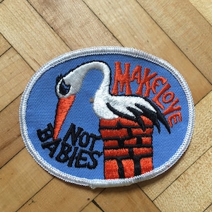 May include: A blue and white embroidered patch featuring a white stork carrying a brick with the text "Make Love Not Babies" in red and black lettering.