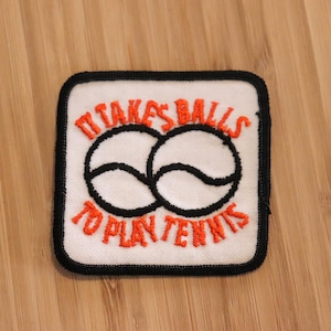 May include: Square patch with the text "IT TAKES BALLS TO PLAY TENNIS" in orange embroidery. The patch features two tennis balls and a black border on a white background.