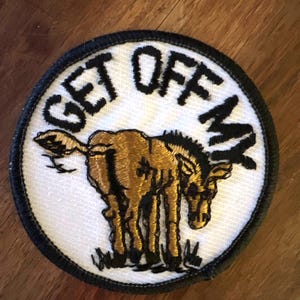 May include: A white, round embroidered patch with a black border. The patch features a gold donkey with the text "GET OFF MY" in black.