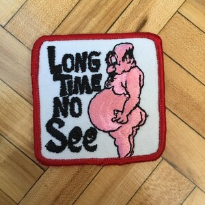 May include: Square patch with a white background and a red border. The patch features the text "LONG TIME NO SEE" in black, along with a pink cartoon figure. The figure has a large belly and is standing.