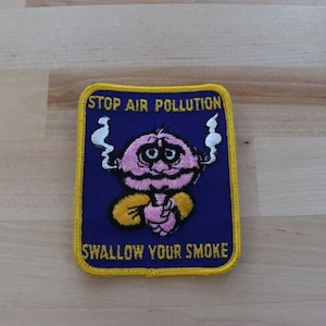 May include: A blue and yellow embroidered patch with a cartoon character smoking a cigarette. The text on the patch reads "Stop Air Pollution Swallow Your Smoke".