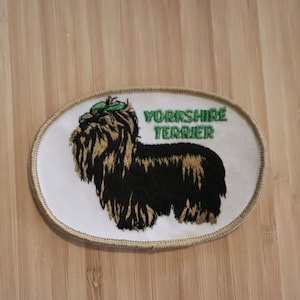 Könnte beinhalten: Gestickter Aufnäher mit einem Yorkshire Terrier Hund mit einer grünen Schleife. Der Aufnäher ist oval geformt mit einem weißen Hintergrund und goldenen Nähten. Der Text "Yorkshire Terrier" ist in grün gestickt.