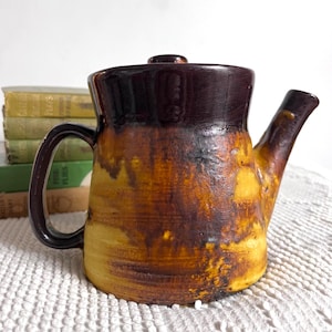 May include: A brown and yellow ceramic teapot with a dark brown lid and handle. The teapot has a mottled glaze and a rustic look.