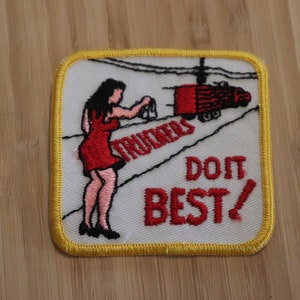 May include: A white embroidered patch with a yellow border. The patch features a cartoon woman in a red dress waving at a red truck with the text "Truckers Do It Best!"