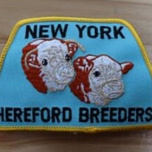 May include: A blue and yellow embroidered patch with the text "NEW YORK HEREFORD BREEDERS" and two white and brown Hereford cattle heads.