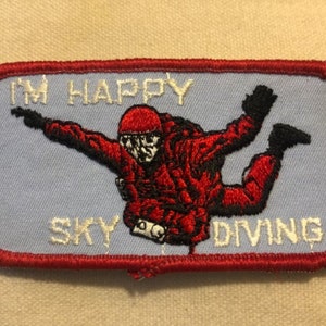 May include: Red, white and blue embroidered patch featuring a skydiver in a red jumpsuit and the text "I'm Happy Sky Diving".