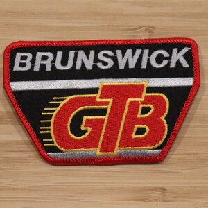 May include: Embroidered patch with the word "BRUNSWICK" in white letters on a black background. Below, the letters "GTB" are in red with yellow accents. The patch has a red border and is on a wooden surface.
