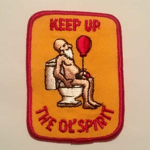 May include: A yellow embroidered patch with red stitching. The patch features a cartoon image of a person sitting on a toilet holding a red balloon. The text on the patch reads "Keep up the good spirit."