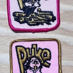 May include: Two pink and white embroidered patches with gold lettering that reads "Duke". Each patch features a cartoon character with a cigar in its mouth.