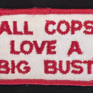 May include: Embroidered patch with a white background and a red border. The patch features the text "ALL COPS LOVE A BIG BUST" in red, stitched lettering. The patch is rectangular.