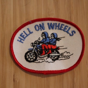 Peut inclure: Écusson brodé blanc avec des coutures rouges. L'écusson représente une moto avec deux motards et le texte "HELL ON WHEELS".