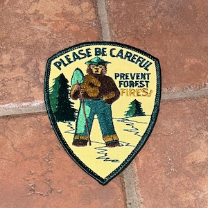 Peut inclure: Écusson brodé représentant Smokey Bear tenant une pelle, avec le texte « PLEASE BE CAREFUL PREVENT FOREST FIRES ! ». L'écusson a un fond jaune avec des détails verts et marron, et une bordure noire. L'écusson est en forme de bouclier.