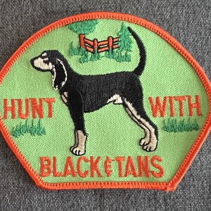 May include: An embroidered patch featuring a black and tan hound dog, with the words "Hunt with Black & Tans" in green and orange.