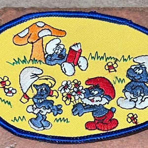 May include: Embroidered patch featuring Smurfs in a whimsical scene. The patch has a yellow background with a blue border. The Smurfs are depicted with blue skin, white hats, and red or white clothing, interacting near a mushroom.