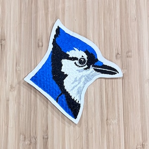 May include: Embroidered blue jay patch with a white and black head and a blue body. The patch is on a white background.