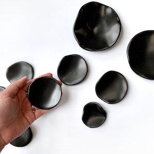 Puede incluir: Colecci&oacute;n de platos de cer&aacute;mica negra con formas org&aacute;nicas e irregulares. Un plato se sostiene en la mano, mientras que los dem&aacute;s est&aacute;n dispuestos sobre una superficie blanca. Los platos var&iacute;an en tama&ntilde;o y tienen un acabado mate.