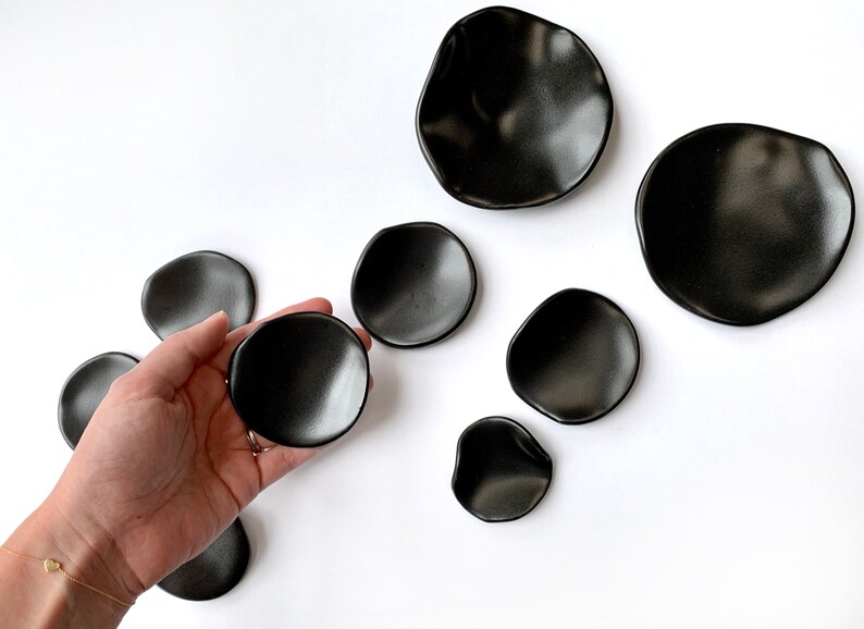 Puede incluir: Colecci&oacute;n de platos de cer&aacute;mica negra con formas org&aacute;nicas e irregulares. Un plato se sostiene en la mano, mientras que los dem&aacute;s est&aacute;n dispuestos sobre una superficie blanca. Los platos var&iacute;an en tama&ntilde;o y tienen un acabado mate.