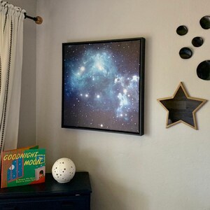 Puede incluir: Una impresi&oacute;n enmarcada de una nebulosa en tonos azules y blancos cuelga de una pared gris. Tambi&eacute;n hay una estrella decorativa de madera y una decoraci&oacute;n de pared circular negra. Una copia de "Goodnight Moon" y una esfera moteada se encuentran en una c&oacute;moda azul oscuro.