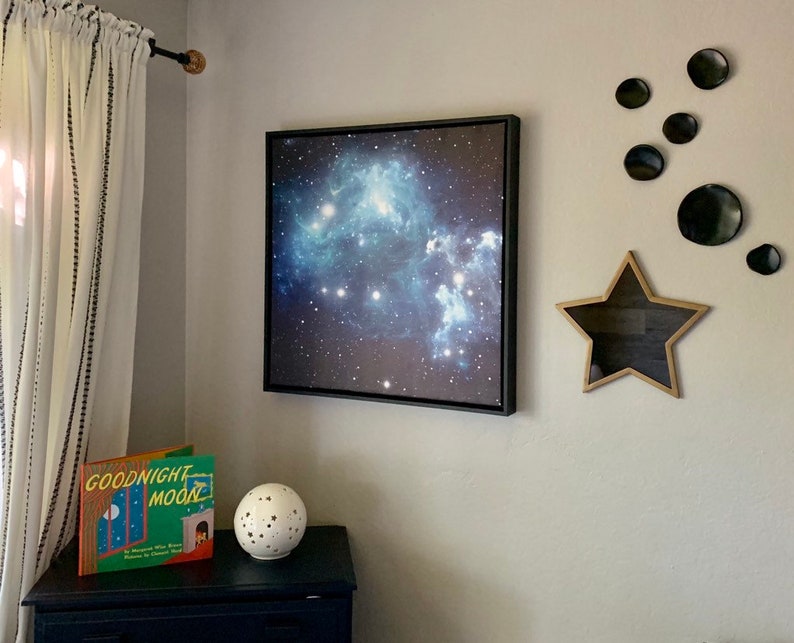 Puede incluir: Una impresi&oacute;n enmarcada de una nebulosa en tonos azules y blancos cuelga de una pared gris. Tambi&eacute;n hay una estrella decorativa de madera y una decoraci&oacute;n de pared circular negra. Una copia de "Goodnight Moon" y una esfera moteada se encuentran en una c&oacute;moda azul oscuro.