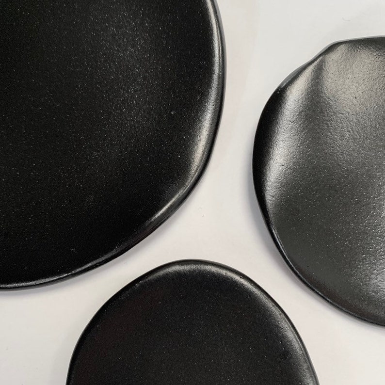 Puede incluir: Primer plano de tres platos de cer&aacute;mica negros de formas irregulares. Los platos tienen un acabado mate y est&aacute;n dispuestos sobre un fondo blanco. Los platos son probablemente para servir comida.