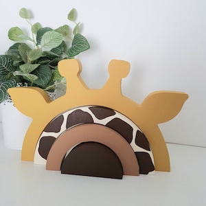 May include: A wooden giraffe stacking toy. The giraffe's head is mustard yellow, with a rainbow-shaped body in shades of brown and white. The toy is designed for children and is made of wood.