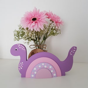 May include: A purple wooden dinosaur toy with a rainbow design. The dinosaur has white polka dots and a curved rainbow with pink and white polka dots. A bouquet of pink flowers sits in a glass jar behind the toy.
