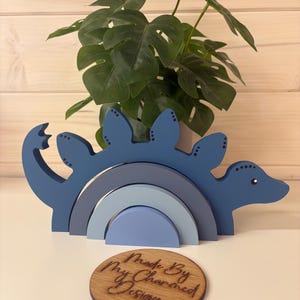 May include: A blue wooden dinosaur toy with a rainbow-shaped base. The dinosaur has a long neck and tail, with a series of blue arches. A wooden disc with the text "Made By My Charmed Design" is in front of the toy.