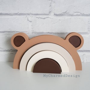 May include: A wooden rainbow stacking toy with brown, beige, and white arches. The toy has two brown bear ears on the top arch.