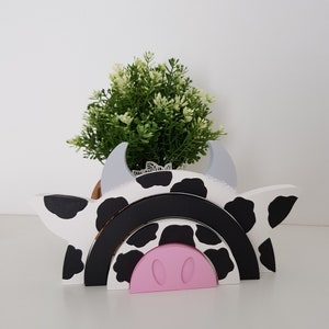 May include: A wooden cow-themed rainbow stacking toy with black and white spots. The toy is made of three arches, with a pink pig nose at the bottom.
