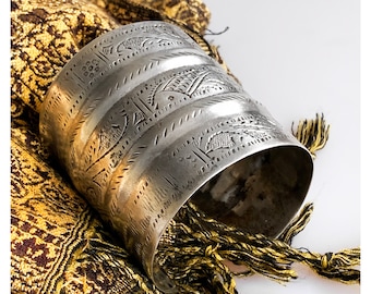 May include: A silver bracelet with an intricate embossed design. The bracelet is wide and has a rounded shape. It is lying on a gold and brown fabric with tassels.