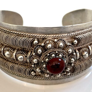 Può includere: Braccialetto rigido in argento con dettagli intrecciati intricati e una grande pietra preziosa rossa al centro.