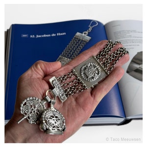 Puede incluir: Un brazalete de cadena de plata con un colgante cuadrado que presenta un diseño detallado. El brazalete tiene un cierre decorativo con dos abalorios colgantes. El brazalete se sostiene en una mano, con un libro en el fondo.