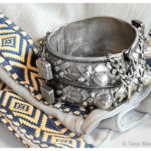 Könnte beinhalten: Ein silberfarbenes Armreif-Armband mit aufwendigen floralen und geometrischen Mustern. Das Armband verfügt über einen Klappverschluss und ist mit dekorativen Elementen wie Ketten und Nieten verziert. Es liegt auf einem blau-gelb gemusterten Textil.