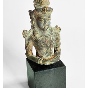 Op de afbeelding: Een bronzen beeld van een godheid met een kroon, die sieraden draagt en op een zwarte basis zit.