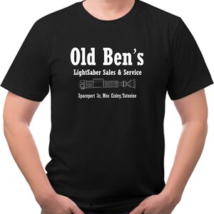 May include: Black t-shirt with white text and a lightsaber graphic. The text reads "Old Ben's Lightsaber Sales & Service" with the location "Spaceport 3c, Mos Eisley, Tatooine" below the graphic. The shirt is a crew neck style.