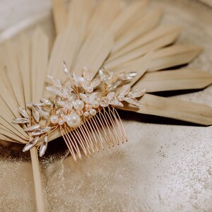 May include: A gold hair comb with a delicate design of leaves and pearls. The comb is lying on a dried palm leaf.