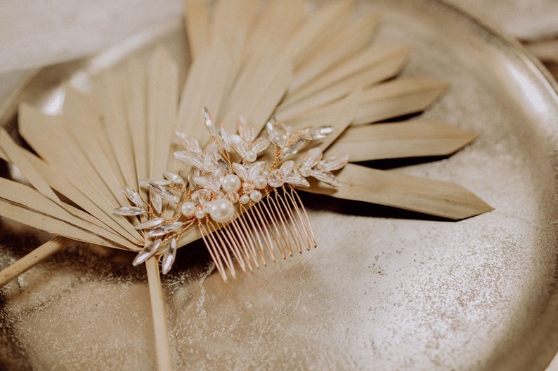 May include: A gold hair comb with a delicate design of leaves and pearls. The comb is lying on a dried palm leaf.