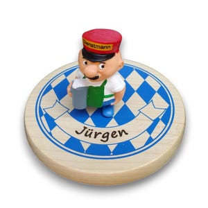 Könnte beinhalten: Ein runder Holzuntersetzer mit blau-weißem Rautenmuster und einem Banner mit dem Namen Jürgen. Eine kleine Figur einer Person mit roter Mütze mit der Aufschrift Dienstmann, weißem T-Shirt, grüner Schürze und blauer Hose hält einen grauen Krug in der Hand.