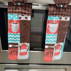 May include: Two brown and white kitchen towels with a coffee cup, coffee pot, and the word "LATTE" printed on them. The towels are hanging on a silver oven door.