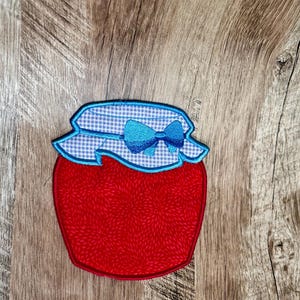 May include: A red jam jar applique with a blue and white gingham lid and a blue bow. The jar has a floral pattern and is outlined in red. The applique is on a wooden surface.