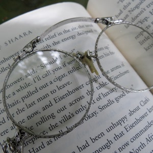 May include: A pair of silver-toned, round, wire-rimmed reading glasses with decorative details on the arms. The glasses are resting on a book with text visible. The text reads "Should I stay?" and "Give the a while. I'll call you if something pulled out a cigar, rolled the last man to leave."