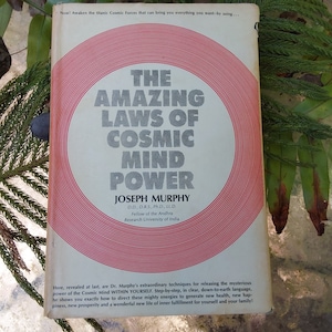 May include: A vintage book cover with a red and white circular design. The title is "The Amazing Laws of Cosmic Mind Power" by Joseph Murphy, D.D., D.R.S., Ph.D., LL.D. Fellow of the Andhra Research University of India.
