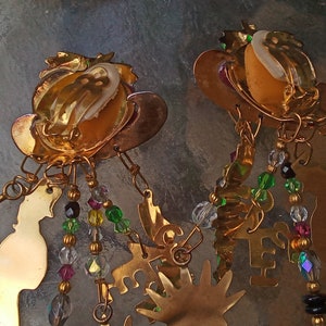 May include: Gold tone earrings with dangling charms and beads. The charms include a small figure, a hand, and a star. The beads are clear, green, and black.