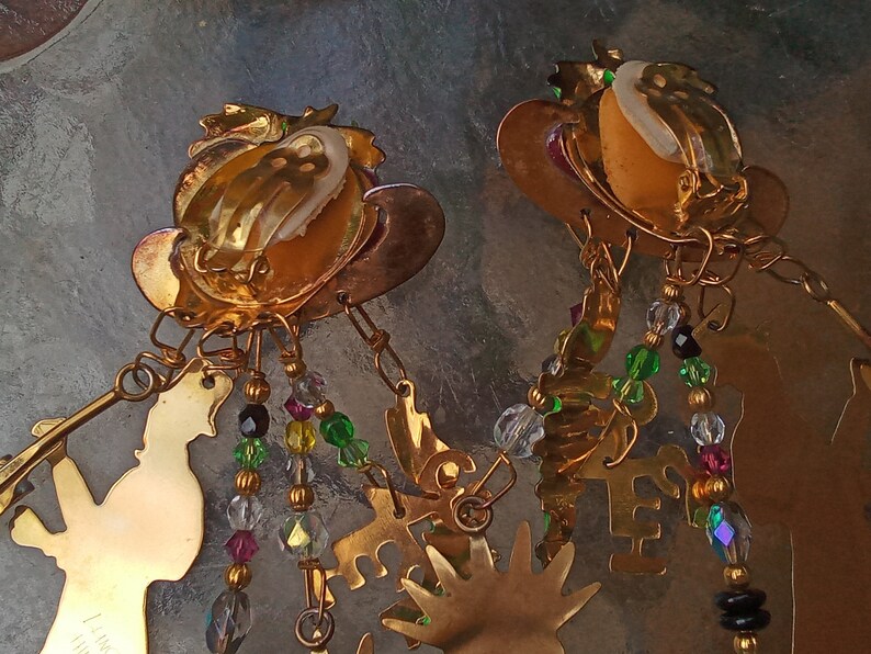 May include: Gold tone earrings with dangling charms and beads. The charms include a small figure, a hand, and a star. The beads are clear, green, and black.